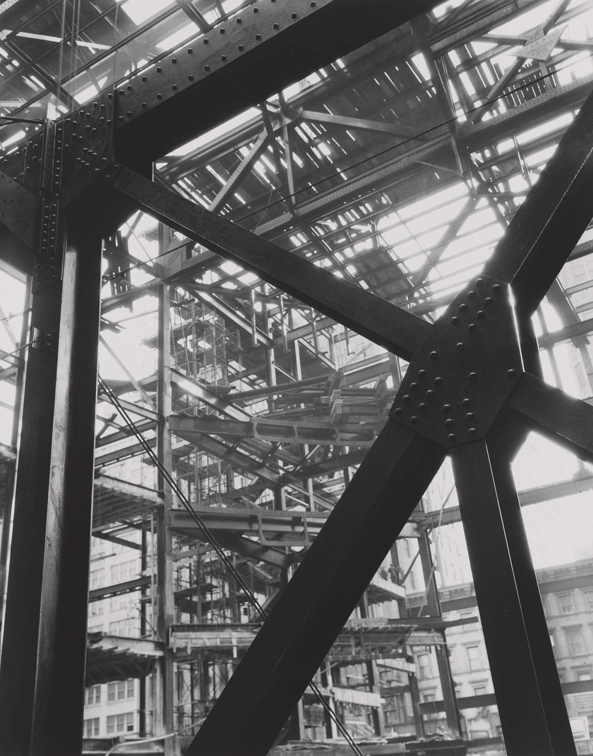 A black and white photograph of a portion of the unfinished building showing exposed steel beams. In the foreground are five beams that meet and are held together with rivets. More of the steel grid of the structure is visible in the background. Wooden planks connect areas of the construction site. Bright sunlight shines through and bleeds around the edges of the beams. A low row of older buildings is barely visible in the background.